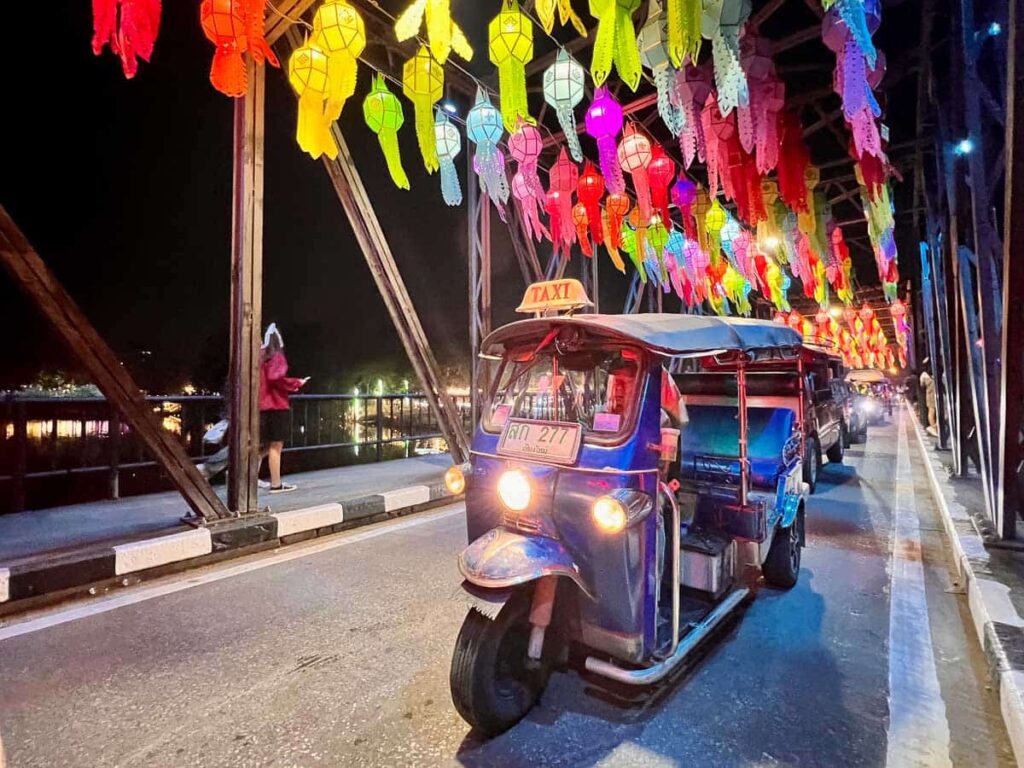 A tuk tuk driving on a bridge with lanterns above.